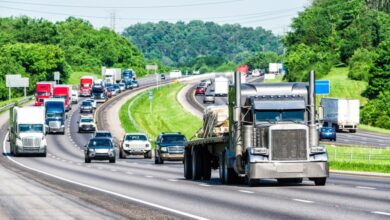 Over a dozen trucks, vans, and cars travel along an interstate highway beside a forest on a cloudy day.