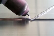 A purple arc flashes between a worn pink TIG welding tip and an aluminum rod held against a metal part.