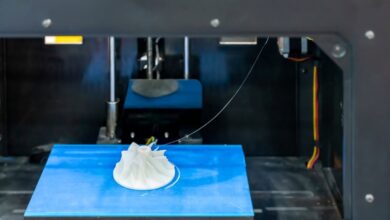 A focus shot of a blue 3D print bed with a white thread of filament connecting from the nozzles to the 3D print.