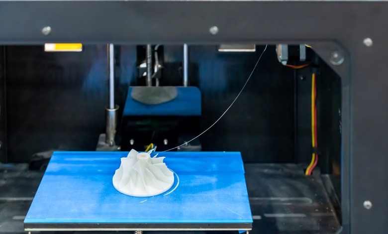 A focus shot of a blue 3D print bed with a white thread of filament connecting from the nozzles to the 3D print.