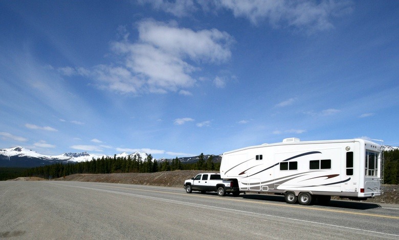 A white truck hauling a large white RV on an open road with trees and snowcapped mountains in the distance.