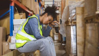 A stressed woman sitting down in a warehouse surrounded by packaged items on various shelves. The area is cramped.