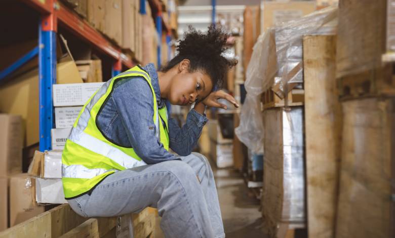 A stressed woman sitting down in a warehouse surrounded by packaged items on various shelves. The area is cramped.
