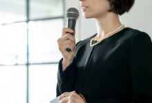A woman in a black funeral dress and white pearls speaks into a microphone while holding a handkerchief in her hand.