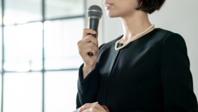 A woman in a black funeral dress and white pearls speaks into a microphone while holding a handkerchief in her hand.