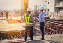 Two contractors in hard hats standing side by side, observing a construction site. One is wearing a safety vest.