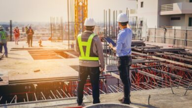 Two contractors in hard hats standing side by side, observing a construction site. One is wearing a safety vest.
