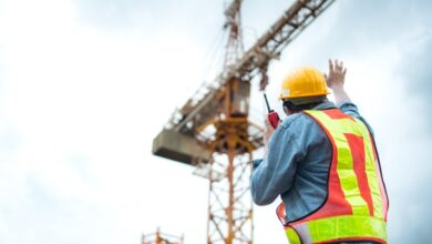 A construction worker in a reflective vest and hard hat holds up his hand to signal to the operator of a crane towering above him.