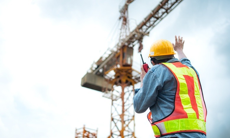 A construction worker in a reflective vest and hard hat holds up his hand to signal to the operator of a crane towering above him.