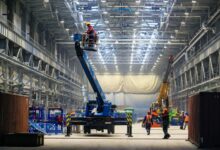 A production warehouse with workers moving around to install new parts. The workers are wearing safety gear.