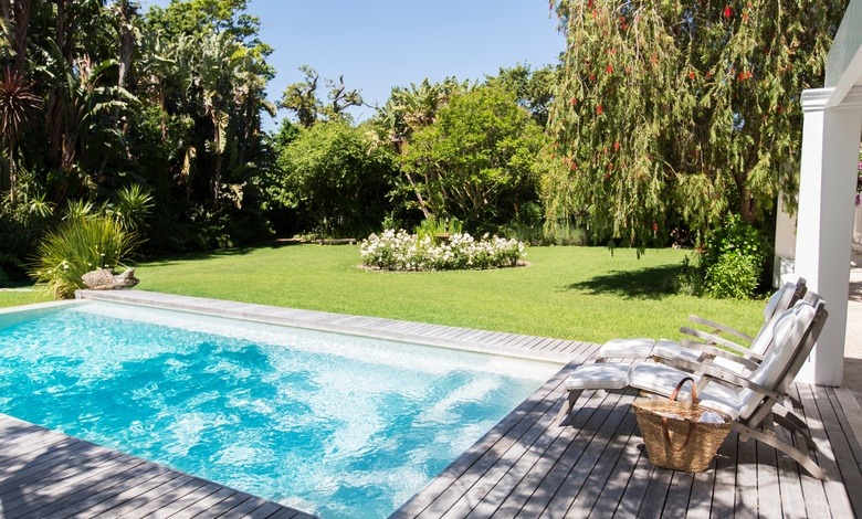 A pool is full of turquoise blue water in a backyard surrounded by gray decking. Two lounge chairs are next to the pool.