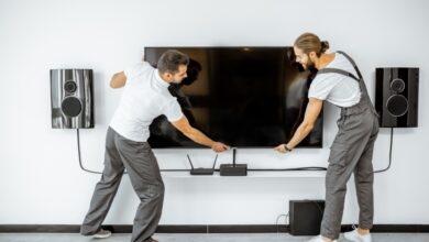 Two professional handymen are installing a large TV monitor and audio system in a modern living room.