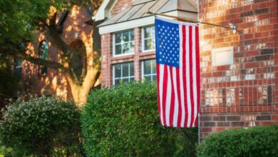An American flag hangs off the end of a wooden flagpole attached to the front of a brick house near bushes.