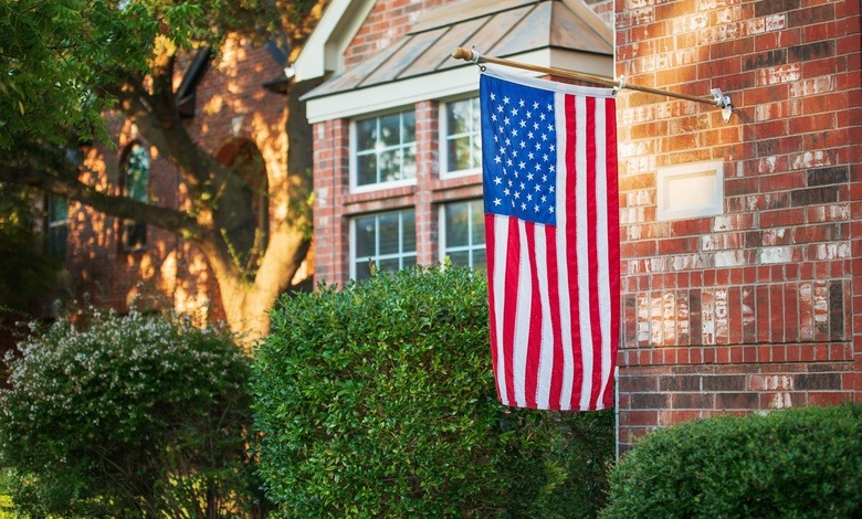 An American flag hangs off the end of a wooden flagpole attached to the front of a brick house near bushes.
