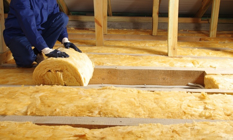 A worker wearing a blue jumpsuit, unrolling a long strip of yellow insulation and placing it on an attic floor.