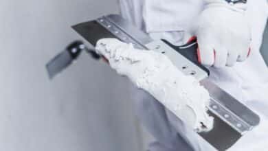 A close-up shot of a worker's gloved hand holding a large taping knife, covered in a large glob of drywall mud.
