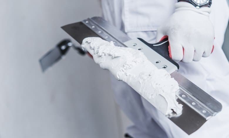 A close-up shot of a worker's gloved hand holding a large taping knife, covered in a large glob of drywall mud.