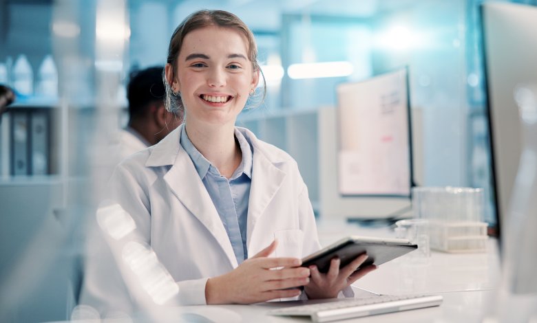 A young smiling woman in a white lab coat sits at a desk in a medical laboratory and holds a tablet.