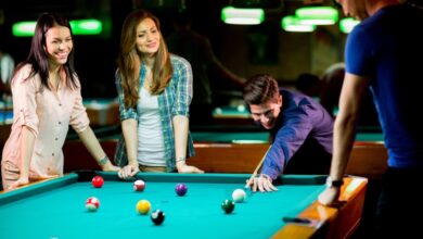 Two young women and two young men are standing around a pool table as one man uses a pool cue to hit a ball.