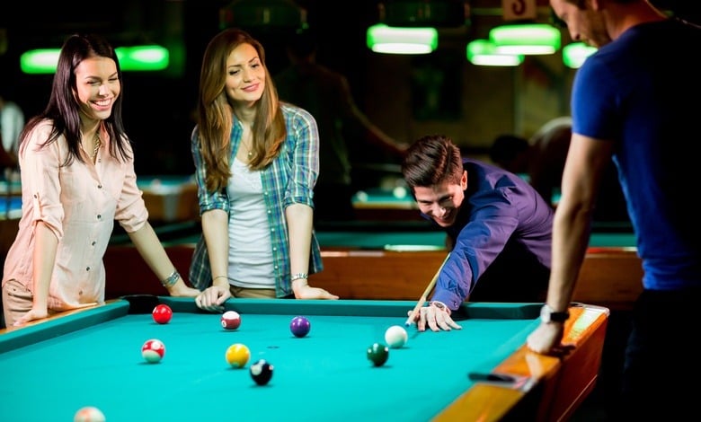 Two young women and two young men are standing around a pool table as one man uses a pool cue to hit a ball.