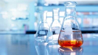 A close up of three glass flasks sitting on a reflective surface in a lab. The frontmost flask has orange liquid in it.