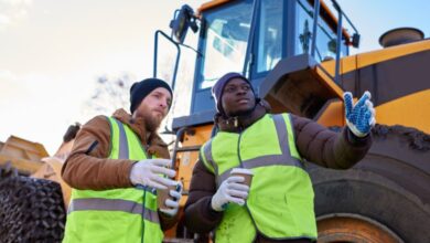 Two workers in coats, beanies, and safety vests stand in front of a piece of construction equipment, holding hot drinks.