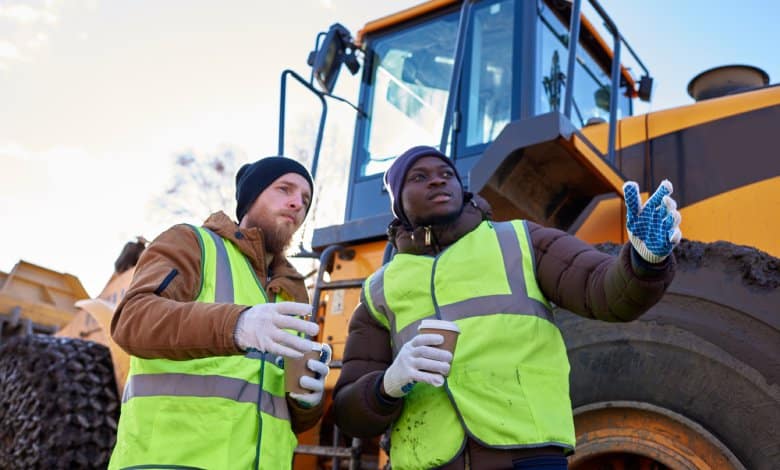 Two workers in coats, beanies, and safety vests stand in front of a piece of construction equipment, holding hot drinks.