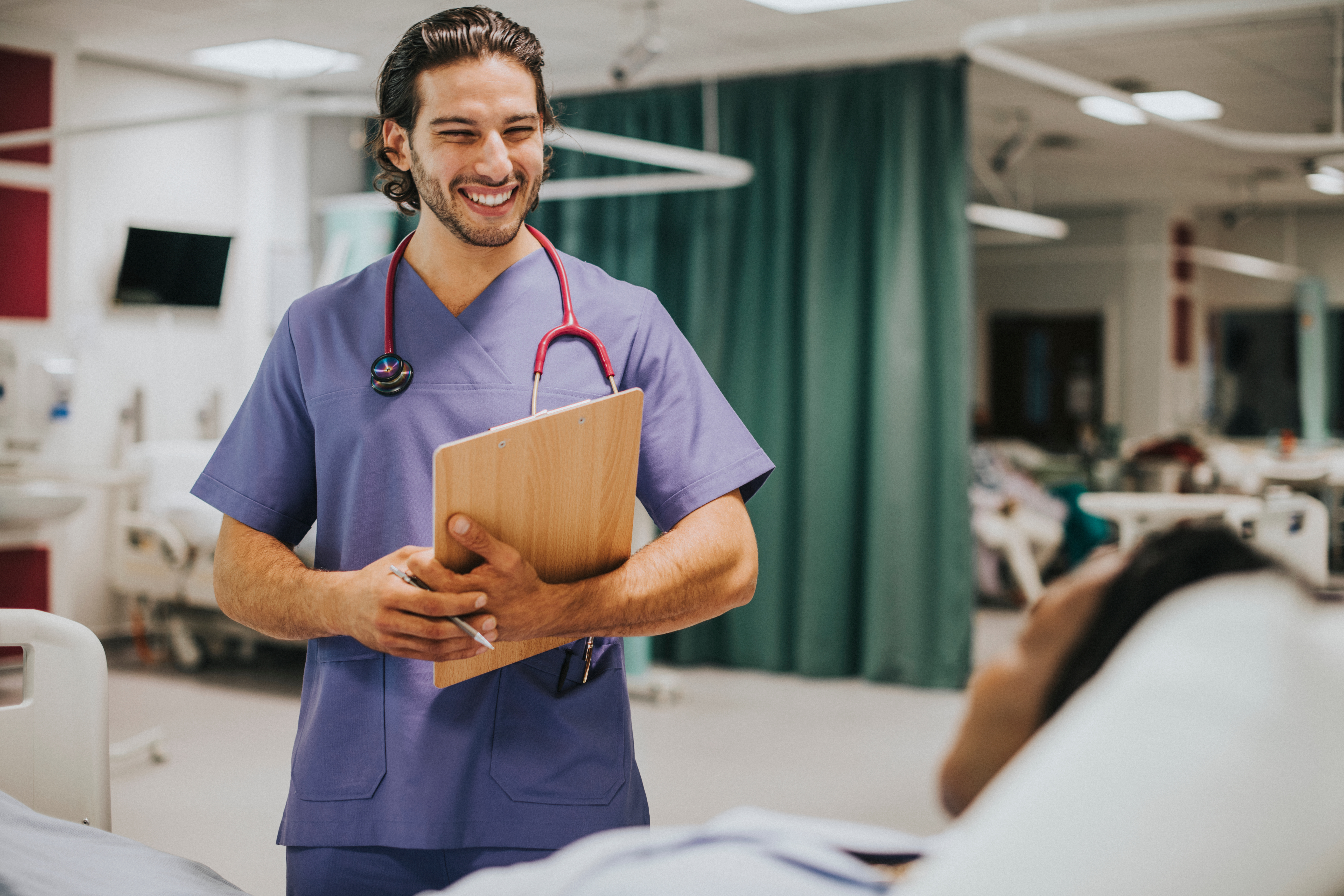 A smiling nurse standing beside a patient in a hospital bed. The nurse is holding a clipboard.