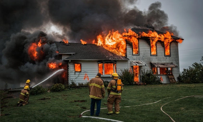 A firefighter uses a hose to put out a massive fire in a home, producing lots of black smoke. Two firefighters stand by.