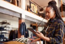 A long-haired young woman in a plaid shirt examining a silver tablet inside a two-story clothing store.