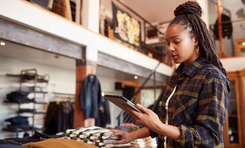 A long-haired young woman in a plaid shirt examining a silver tablet inside a two-story clothing store.