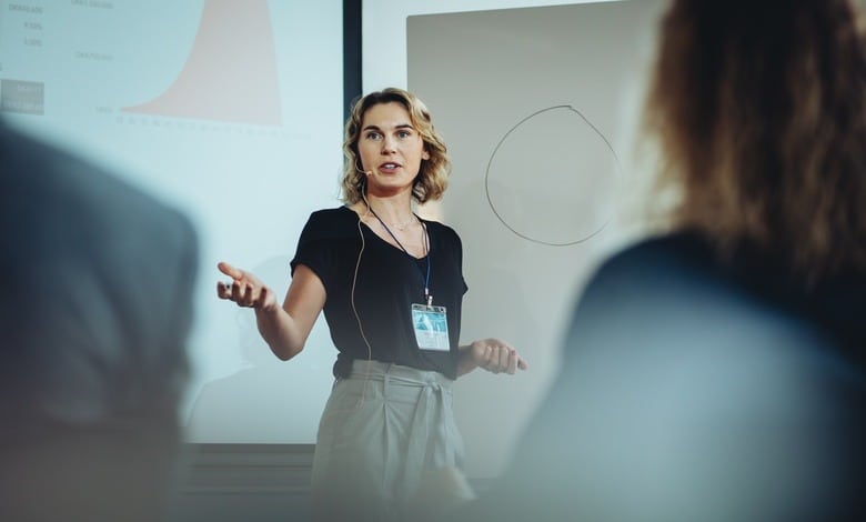 A blonde-haired woman in a black top and gray pants delivers a presentation using a hands-free microphone.