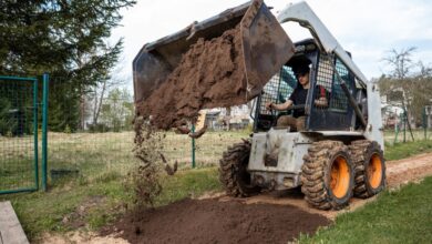 A young man is operating a white skid steer. The skid steer is dumping soil onto a paved dirt pathway.