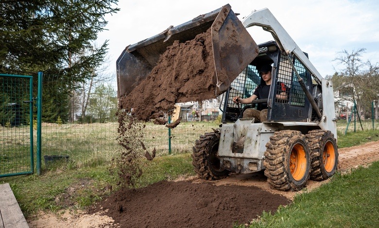 A young man is operating a white skid steer. The skid steer is dumping soil onto a paved dirt pathway.