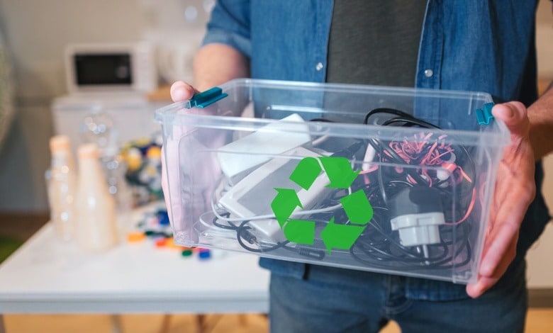 A close-up of a man holding a clear plastic recycling bin that is filled to the top with old electronic devices.