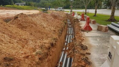 Ground next ot a sidewalk in front of a street dug up to reveal multiple lines of underground utilities.
