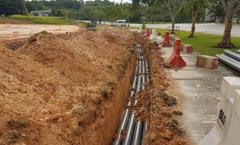 Ground next ot a sidewalk in front of a street dug up to reveal multiple lines of underground utilities.