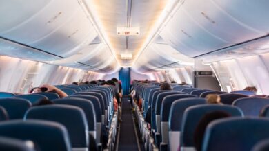 An airplane cabin interior with passengers seated in three blue seats on each side of the narrow aisle.
