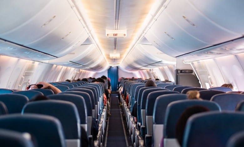 An airplane cabin interior with passengers seated in three blue seats on each side of the narrow aisle.