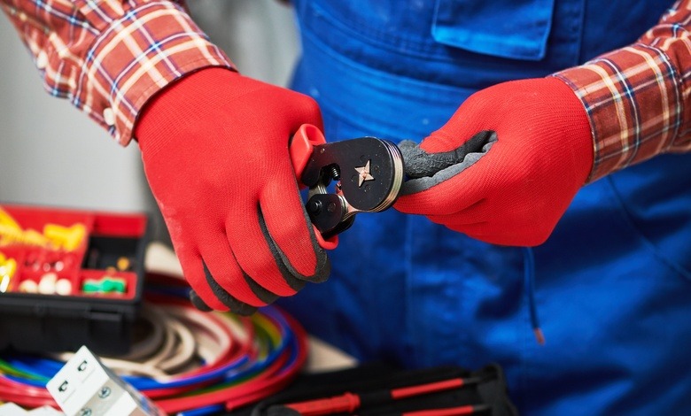 A close-up of a man's hands with red work gloves using a hand crimping tool to crimp an electrical connection.