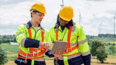 Two construction zone workers dressed in yellow reflective vests and yellow hard hats stand together discussing plans.