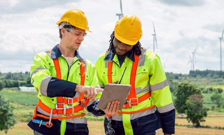 Two construction zone workers dressed in yellow reflective vests and yellow hard hats stand together discussing plans.