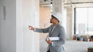A person wearing a business suit and a hard hat is holding a rolled-up blueprint while inspecting a construction site.