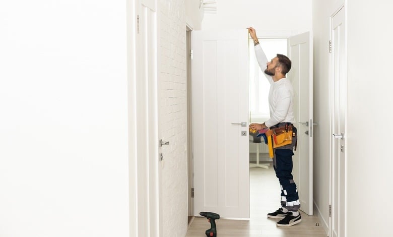 A handyman wearing a white shirt and a toolbelt installs a white wooden door in a bright, naturally lit hallway.