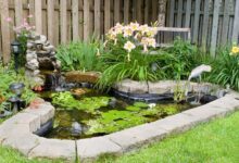 A small pond covered in water lilies with a rock edge, a fake wading bird, a small fountain, and multiple plants.