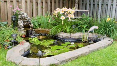 A small pond covered in water lilies with a rock edge, a fake wading bird, a small fountain, and multiple plants.