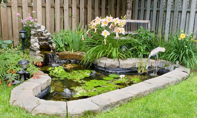 A small pond covered in water lilies with a rock edge, a fake wading bird, a small fountain, and multiple plants.