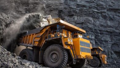 A mining dump truck is loading coal into the back of the vehicle. A wall of black rocks is in the background.