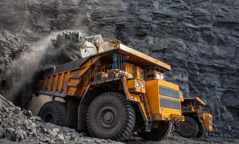 A mining dump truck is loading coal into the back of the vehicle. A wall of black rocks is in the background.