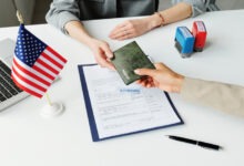 A person hands a passport holder across the table to another person. On the table is an approved form and a U.S. Flag.